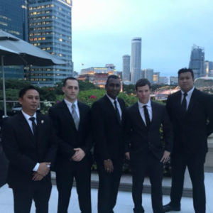 Five smartly presented men in black suits stand at ease in-front of the Singapore skyline